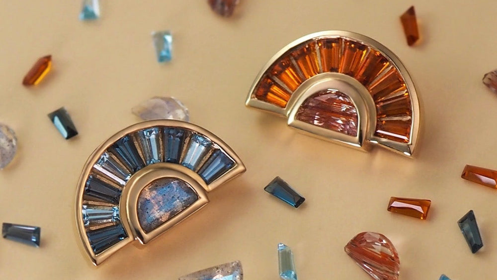 Two BVLA solid 14k gold custom piercing jewellery designs featuring blue and orange baguette-cut gemstones with complementary centre gemstones, displayed on a luxury gemstone styling board.