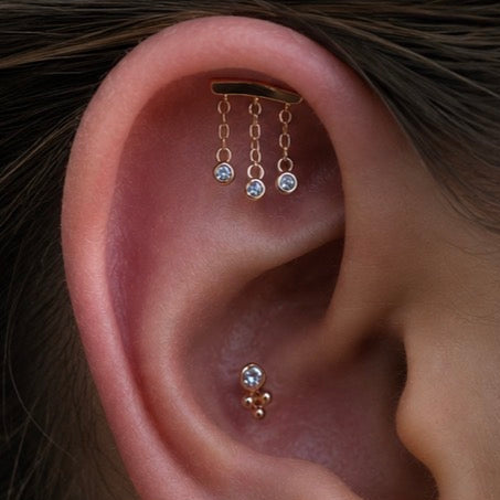Curated ear featuring a Junipurr Delilah hidden helix chain, Tri Bead conch end, bezel-set cubic zirconia cluster, Tri Bead lobe end, CZ prong stud and gold huggie with a gemstone charm, all styled in solid 14k gold.