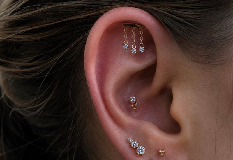 Luxury hidden helix jewellery featuring the Junipurr Delilah chain end, 14k gold bezel-set conch stud, tri bead conch piercing, stacked lobe piercings and gold charm hoop.