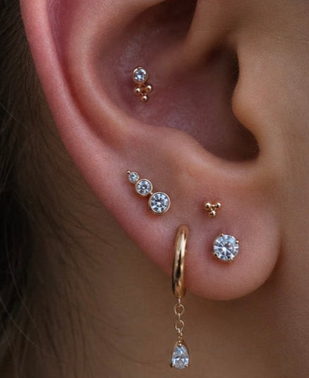 Curated ear styled with Junipurr white CZ jewellery, featuring a CZ tri bead in the conch, a gold tri bead stud, stacked white CZ lobe studs, and a yellow gold huggie hoop with a white CZ chain dangle charm for a luxury ear stack.