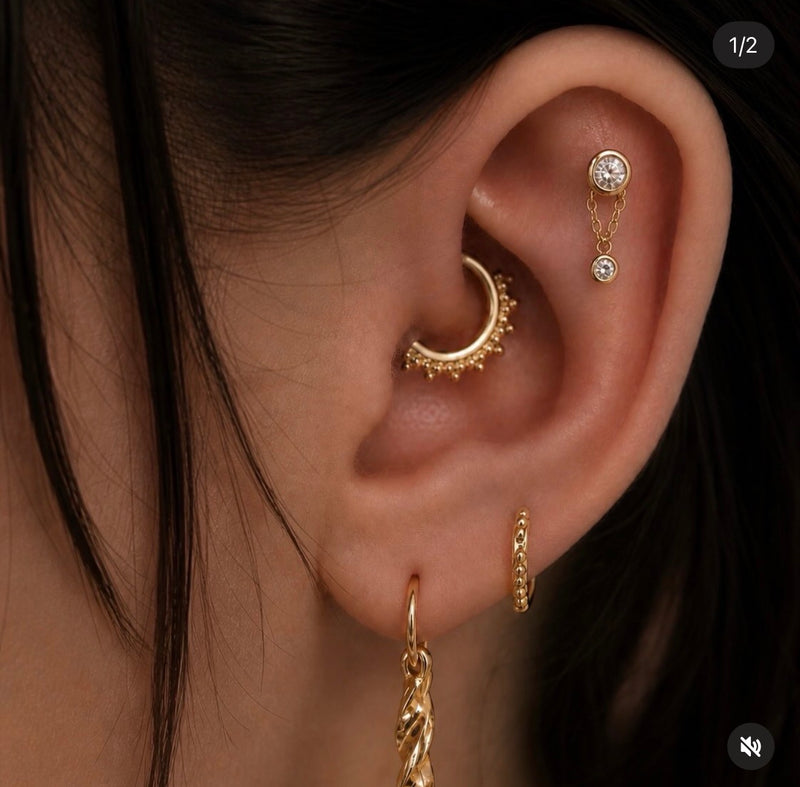 Curated ear featuring a Junipurr white CZ Cora chain dangle in a flat helix piercing, paired with a yellow gold beaded daith clicker and gold huggie hoops for a luxury ear stack.