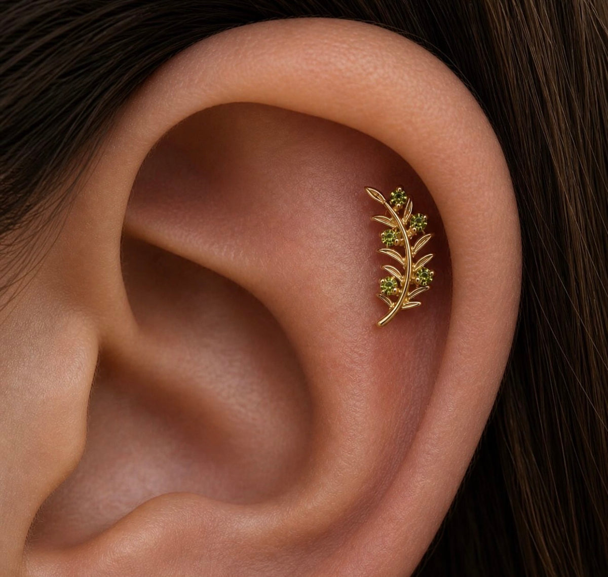 Close-up of a healed helix piercing featuring the Junipurr Olive Branch end in solid 14k yellow gold with green cubic zirconia accents styled along the outer ear.