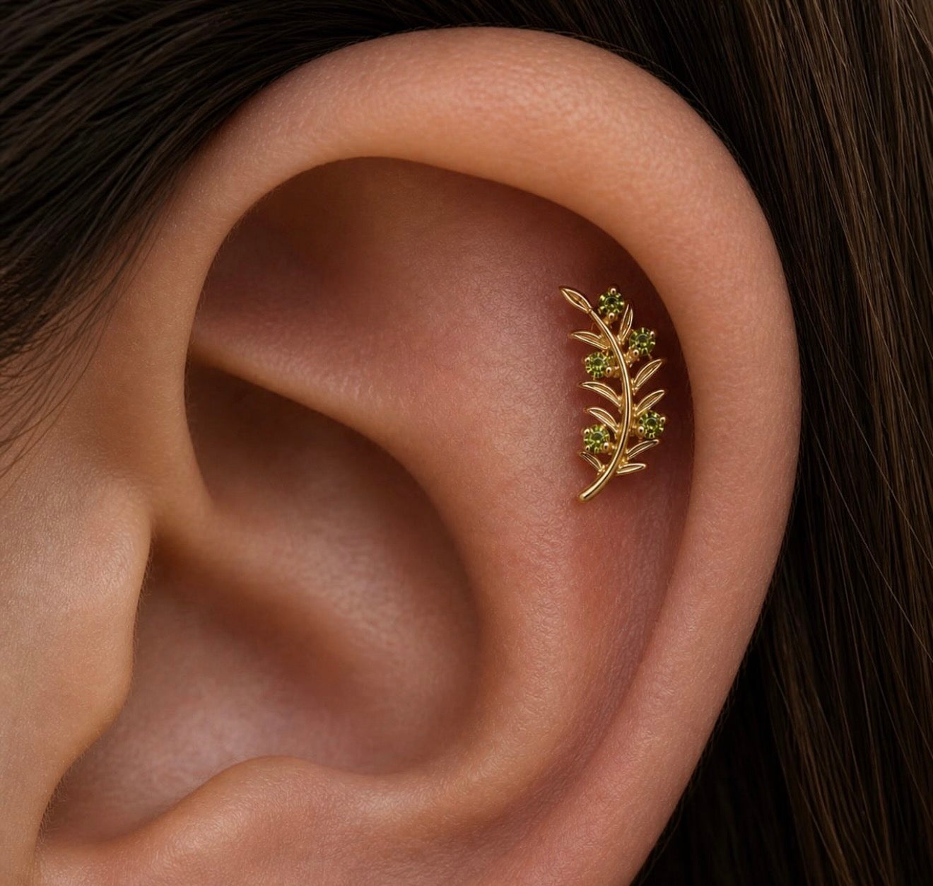 Close-up of a healed helix piercing featuring the Junipurr Olive Branch end in solid 14k yellow gold with green cubic zirconia accents styled along the outer ear.