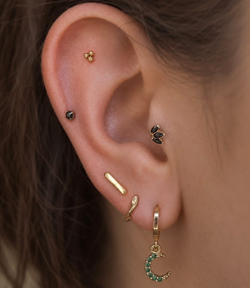 Close-up of a curated ear featuring black CZ marquise tragus jewellery, a black CZ helix stud, a gold tri-bead flat helix stud, minimalist gold lobe jewellery, and a gold crescent moon charm hoop.