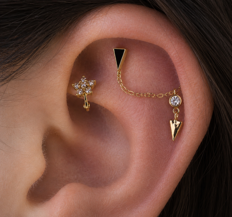 Close-up of a curated ear featuring a luxury gold hidden helix chain with geometric black and gold ends, a gold flower conch ring with white gemstones, and fine 14k gold piercing jewellery.